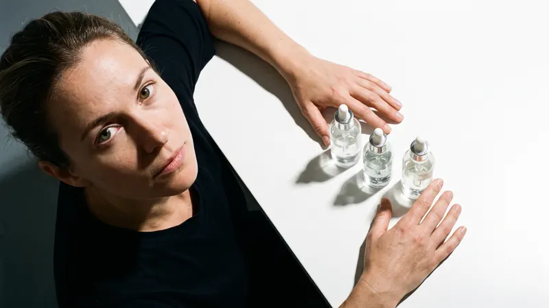 A woman at a white table seen from above, three glass dropper bottles laid out in a row in front of her, lit by hard light from directly overhead casting sharp shadows.