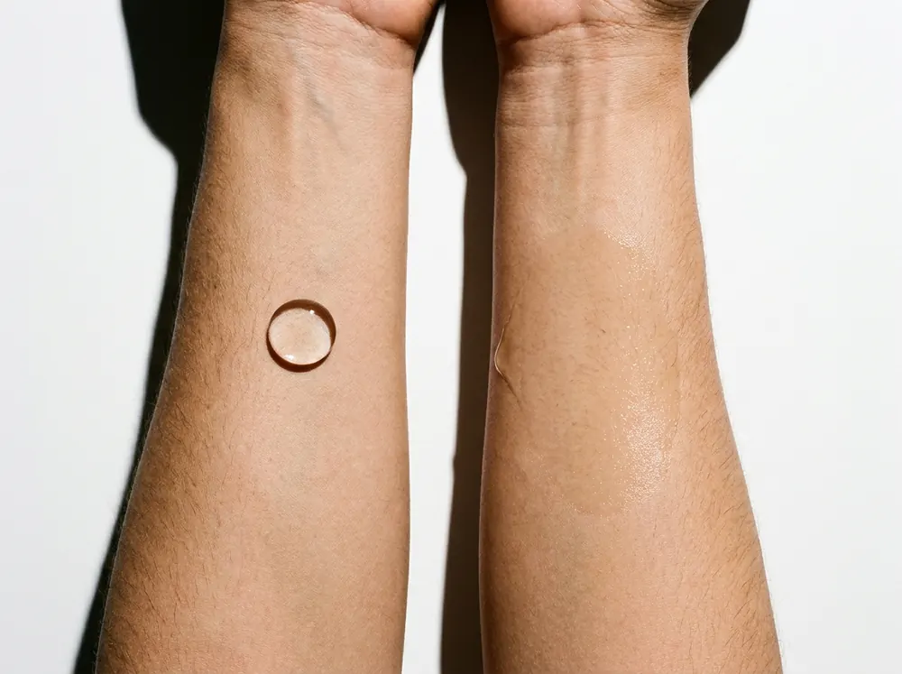 Two forearms side by side on a white surface under hard overhead light, a clear droplet beading up on one and an oilier one spread flat and sunk into the skin on the other.