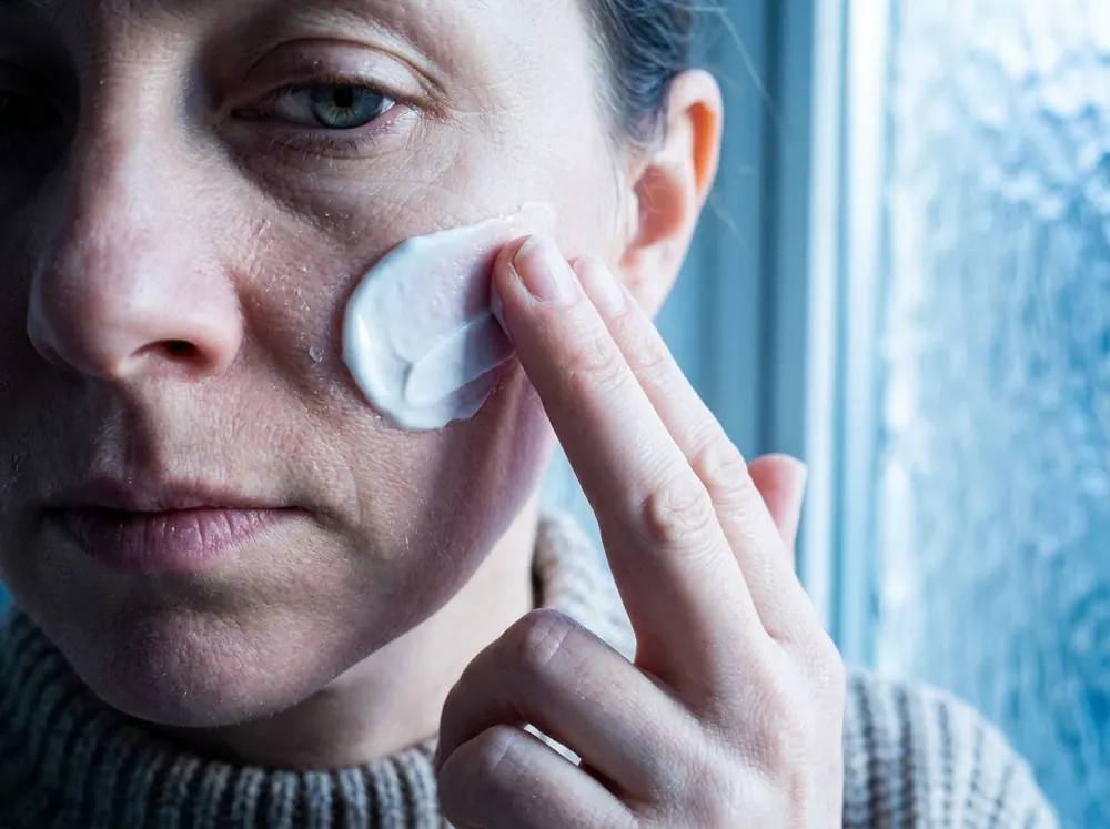 A woman pressing a thick white cream onto her cheek beside a frosted bathroom window in cold blue daylight, the cream visibly sitting on the surface of the skin with dry flaking beside it.