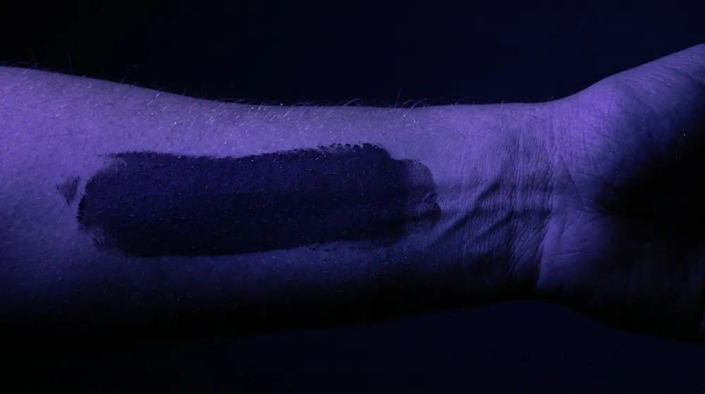 A forearm and wrist lit by violet black light in a dark room, a streak of sunscreen showing as a dark band against the faintly glowing skin.