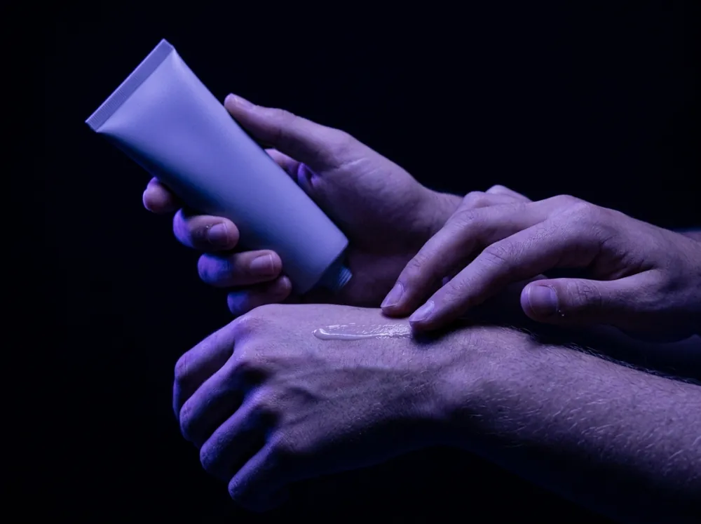 Hands in violet light against a black background, one holding a pale unmarked tube while a thin line of cream is spread on the back of the other hand.