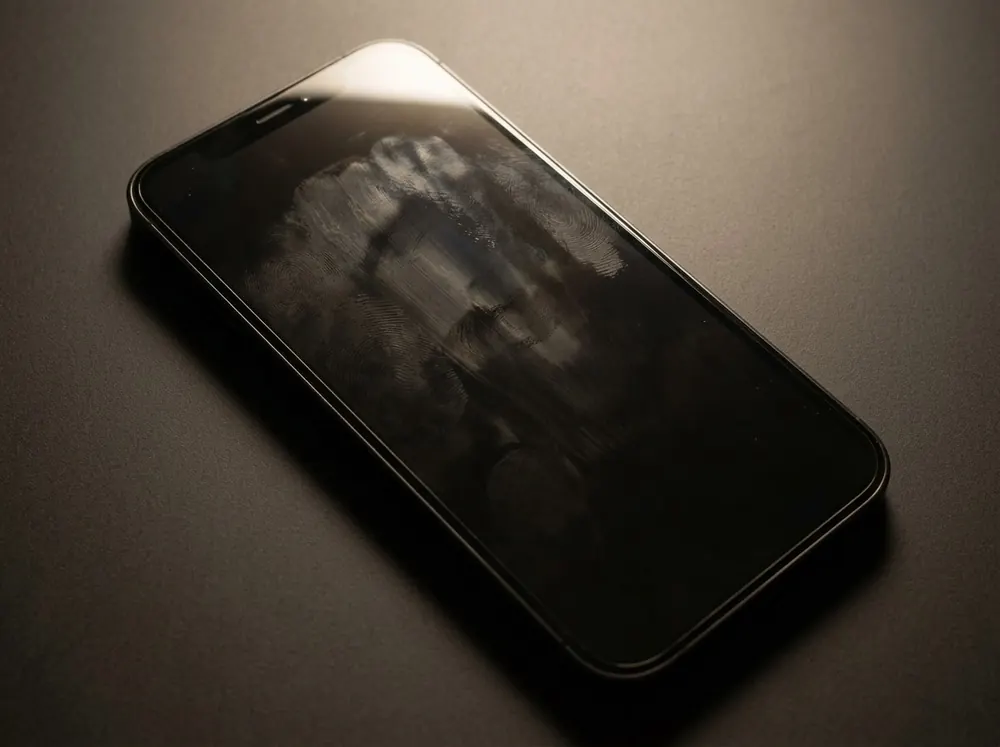 A phone lying screen-up on a dark surface, its switched-off screen lit at a low angle so fingerprint and cheek smudges become visible.