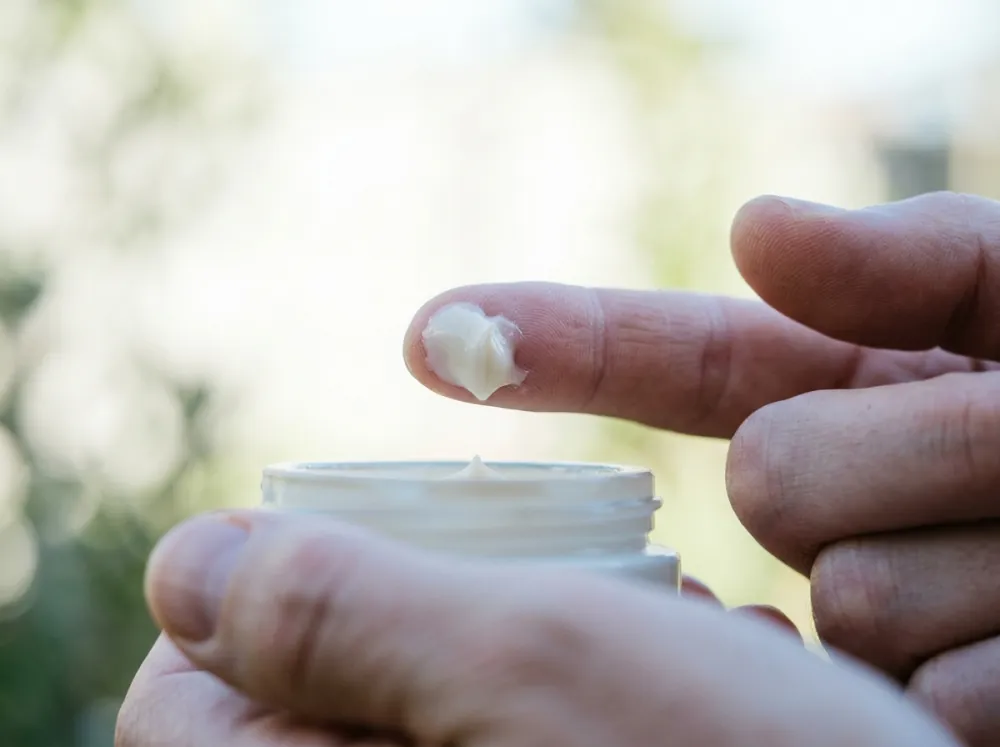 A fingertip holding a small dab of white cream above an open jar, against a bright blurred garden background.