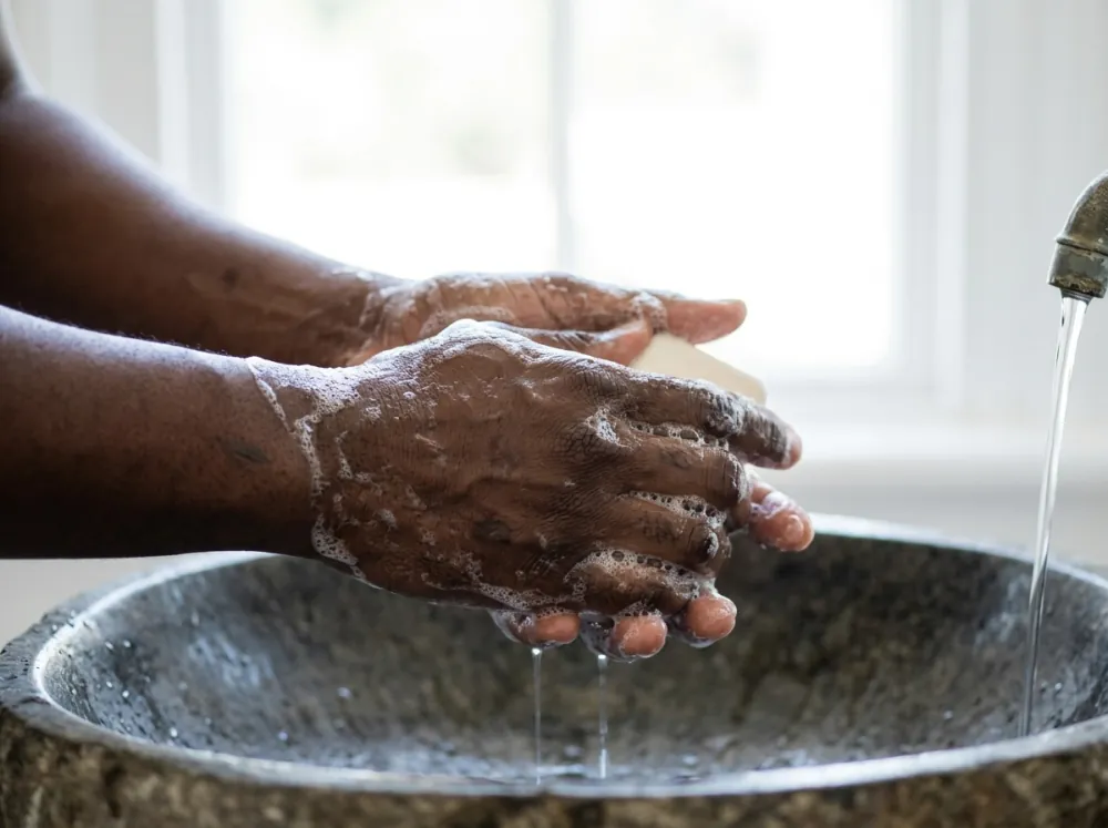 Deep-brown hands lathering a bar of soap over a stone basin, foam on the skin and water running from a tap, with a bright blurred window behind.