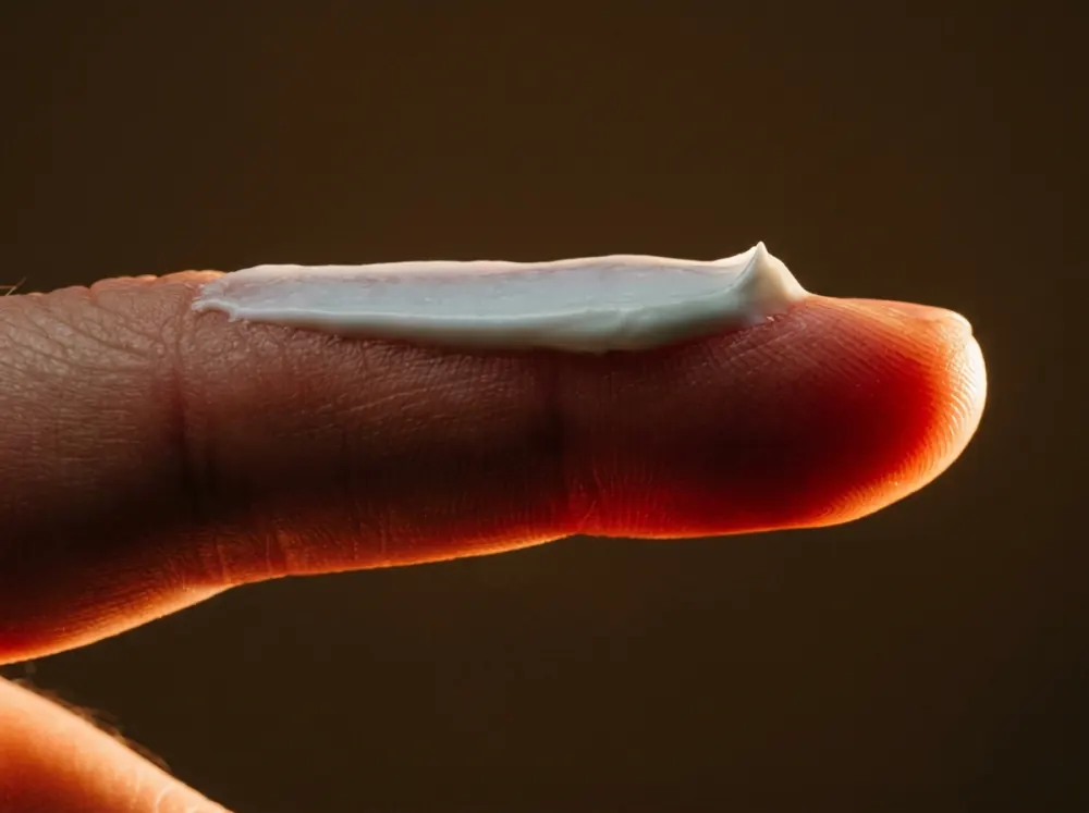The side of an index finger with a smear of opaque white cream lying on top of the skin, while the skin of the fingertip beneath and beside it glows translucent red-orange against a dark brown background.