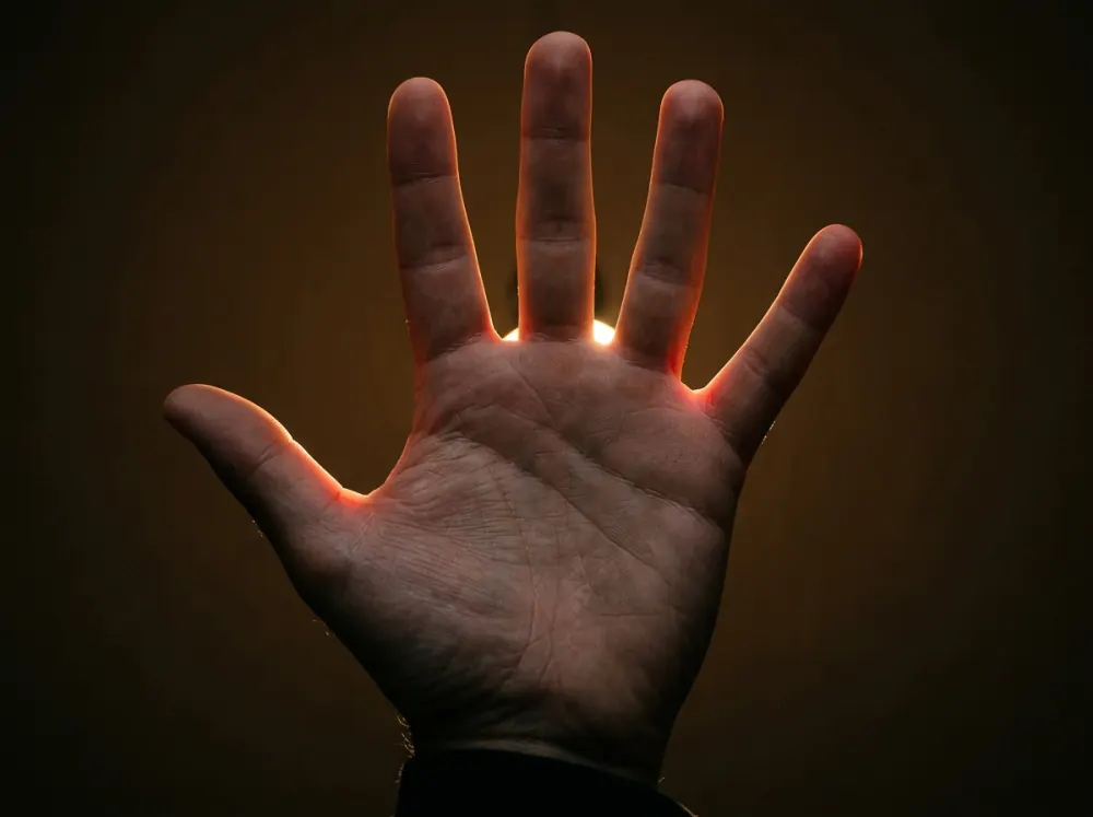 An open hand held up palm-out against a dark background, a bright light behind it showing as a red-orange glow in the thin webbing between the fingers while the palm and fingers stay dark.
