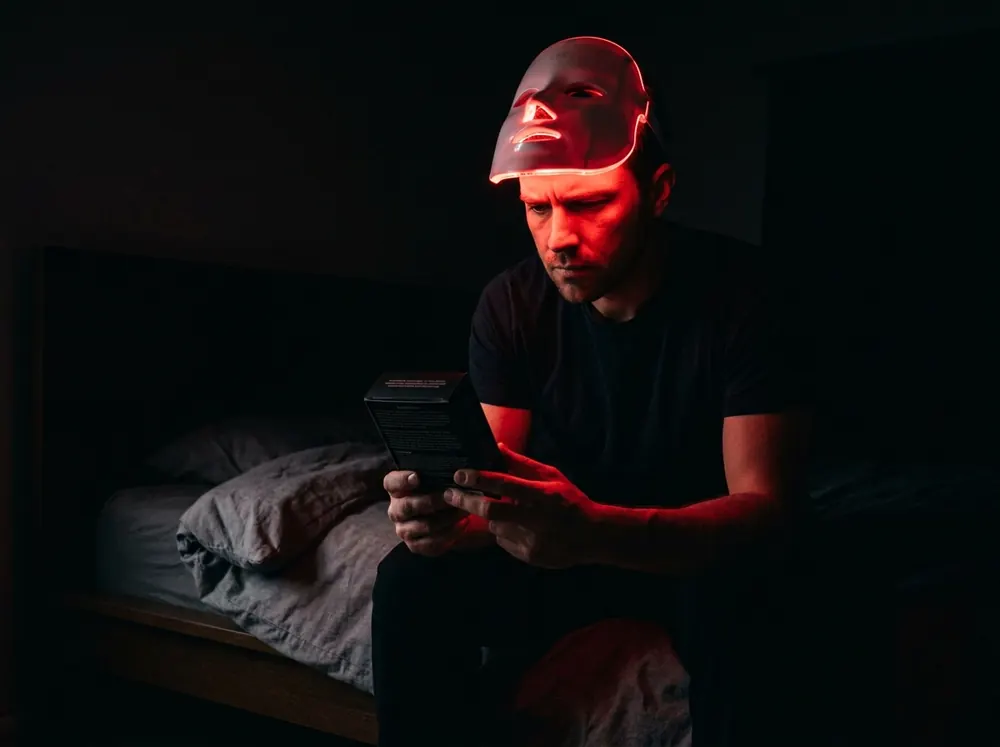 A person sitting on the edge of a bed in a dark room with an LED mask pushed up onto their forehead, reading the small print on the back of the device's box by the mask's red glow.
