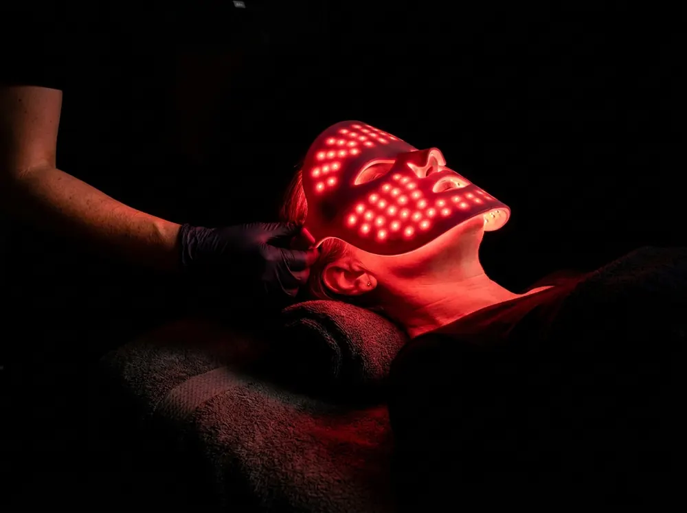 A person lying back on a treatment couch in a dark clinic room while a gloved hand adjusts an LED mask over their face, the mask's red glow the only light in the room.
