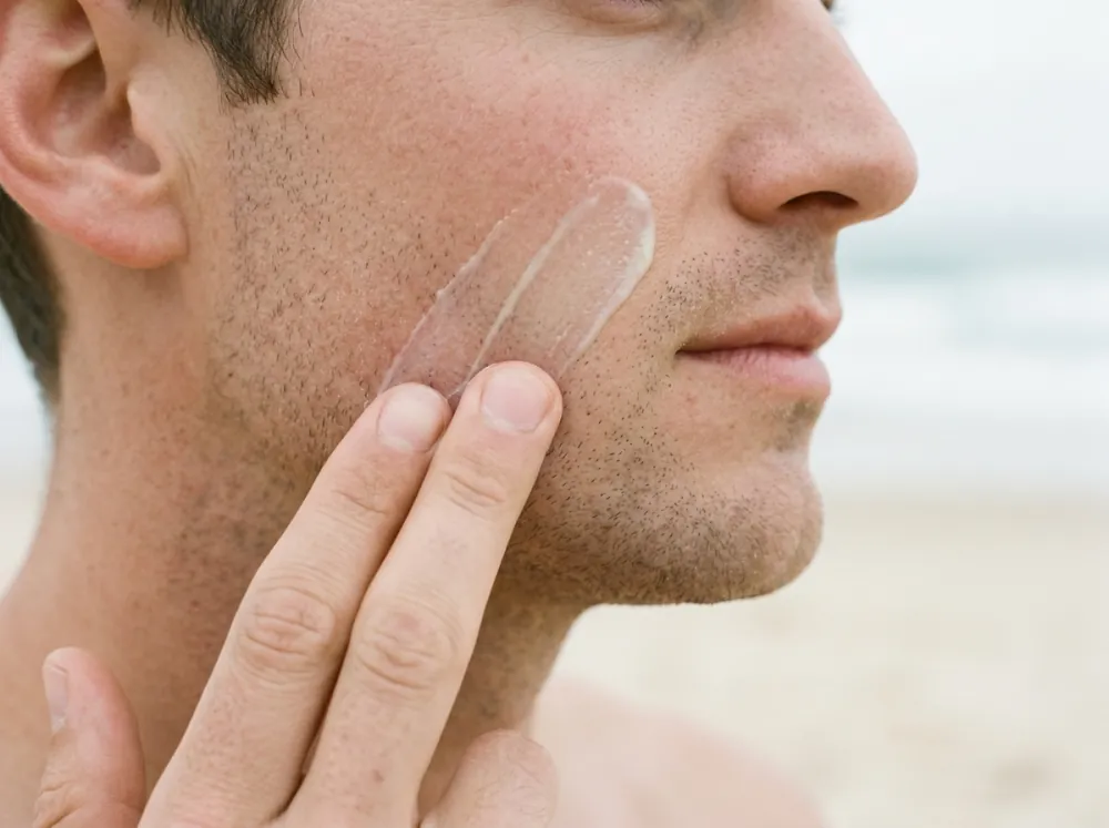 Two fingertips drawing a clear, glossy sunscreen across a man's cheek in profile, leaving a translucent film behind them against a blurred beach background.