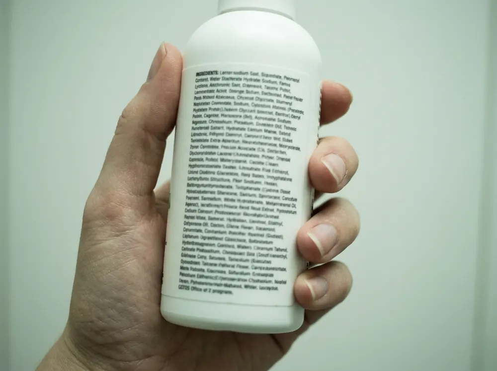 A hand holding a plain white bottle turned to show the ingredient list, the text too small to read in cold overhead light.