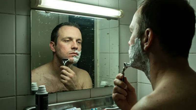 A man at a bathroom mirror under a harsh overhead strip light, half his jaw shaved and the rest still covered in foam, the light casting a flat green tinge across his face.