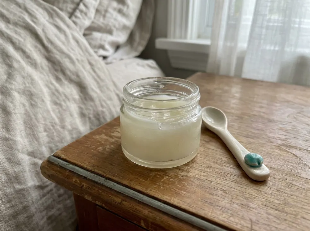 A small jar of clear ointment beside a ceramic spoon on a bedside table