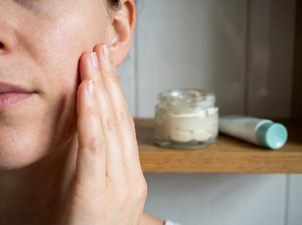 Applying moisturiser to the cheek near a shelf holding a cream jar and a tube