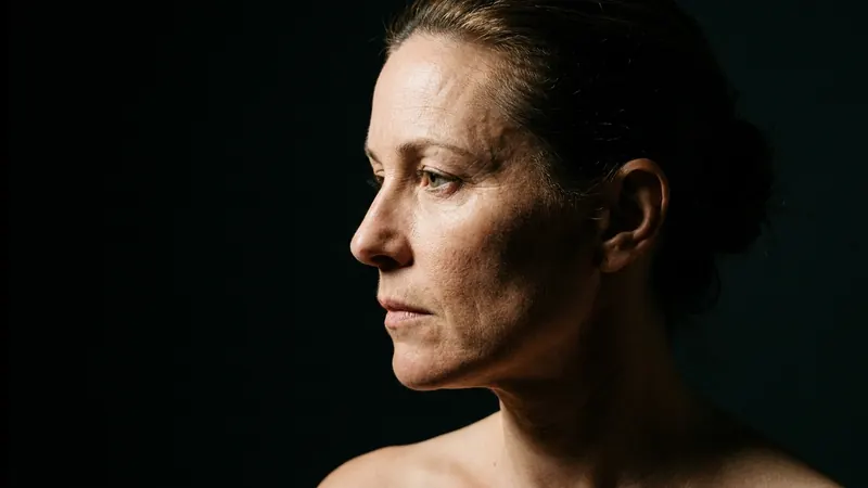 A woman's face in profile against a dark background, lit by a single hard light from the side so the texture of the skin at her temple and jaw stands out in relief.