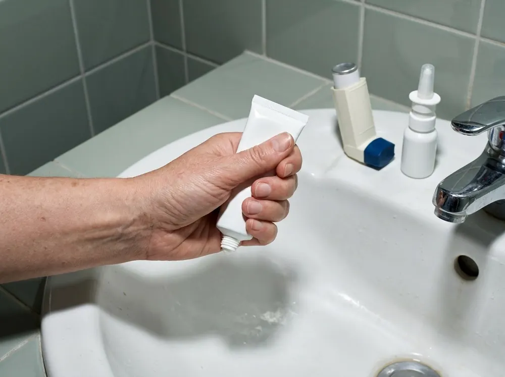 A hand holding a plain, heavily squeezed white cream tube over a bathroom basin, with an unlabelled inhaler and nasal spray bottle standing beside the tap.