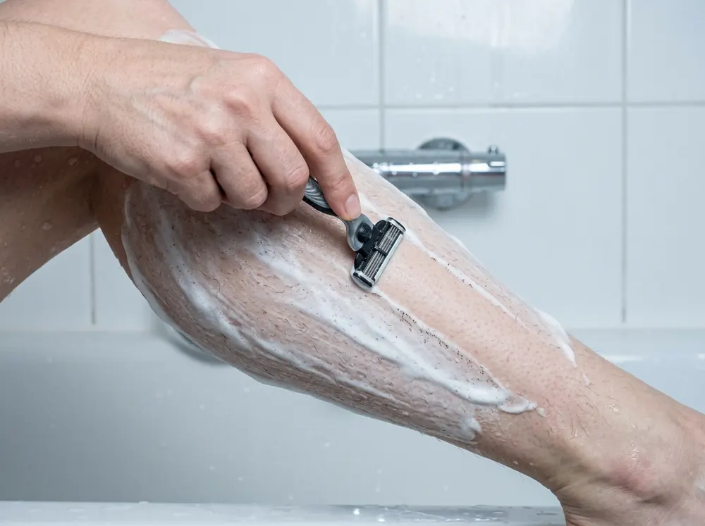 A person shaving a lower leg in the direction of hair growth, blade moving downward over foam, in bright bathroom light.