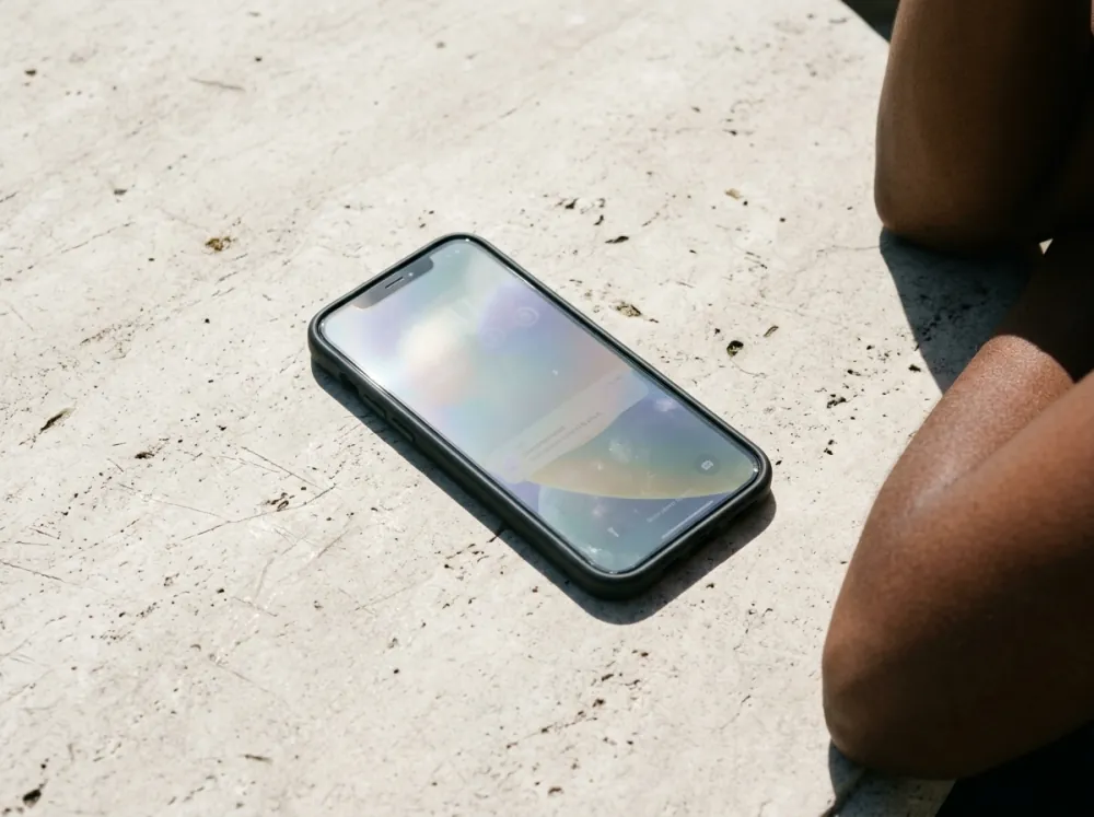 A smartphone in a dark case lying screen-up on a pale stone surface in direct sun, its screen looking pale and washed out, with part of a person's bent arm at the right edge of the frame.