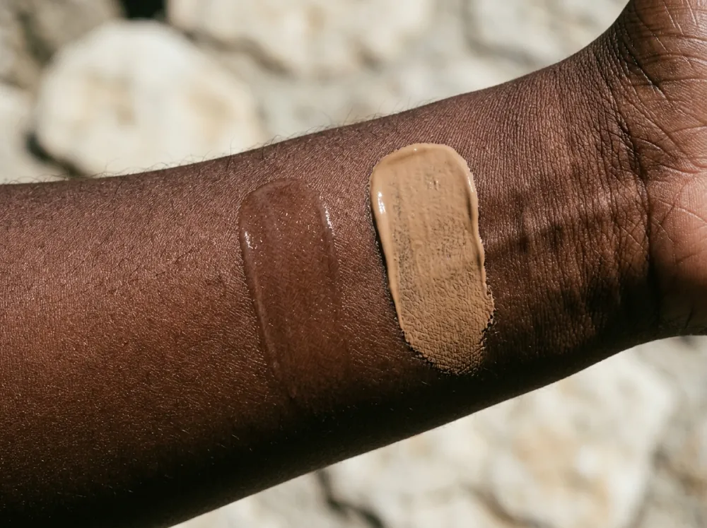 A deep brown forearm in sun with two smears of product side by side, the left a glossy, nearly transparent brown film and the right an opaque beige film with a clear edge.