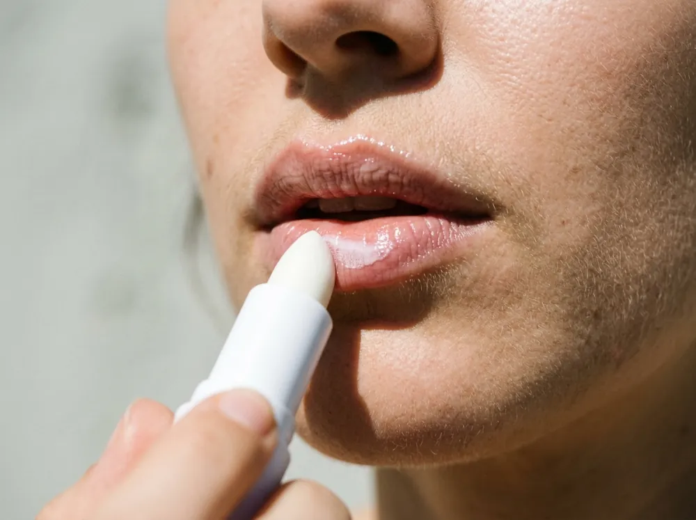 The lower half of a woman's face in hard sun, the tip of a white lip stick touching her lower lip, with a glossy film already on both lips.