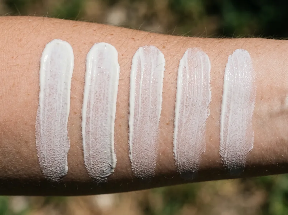 A forearm outdoors with five parallel white stripes of sunscreen side by side, each a slightly different thickness, with bare skin showing between them.