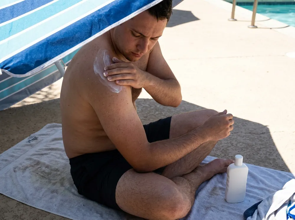 A person reapplying sunscreen to a shoulder in the shade of a beach umbrella, hard sunlight visible beyond the shade line.