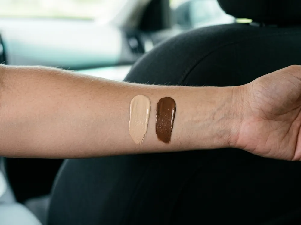 A forearm held in front of a black car seat with two swatches of tinted cream, a light beige and a deep brown, each with a clear edge.