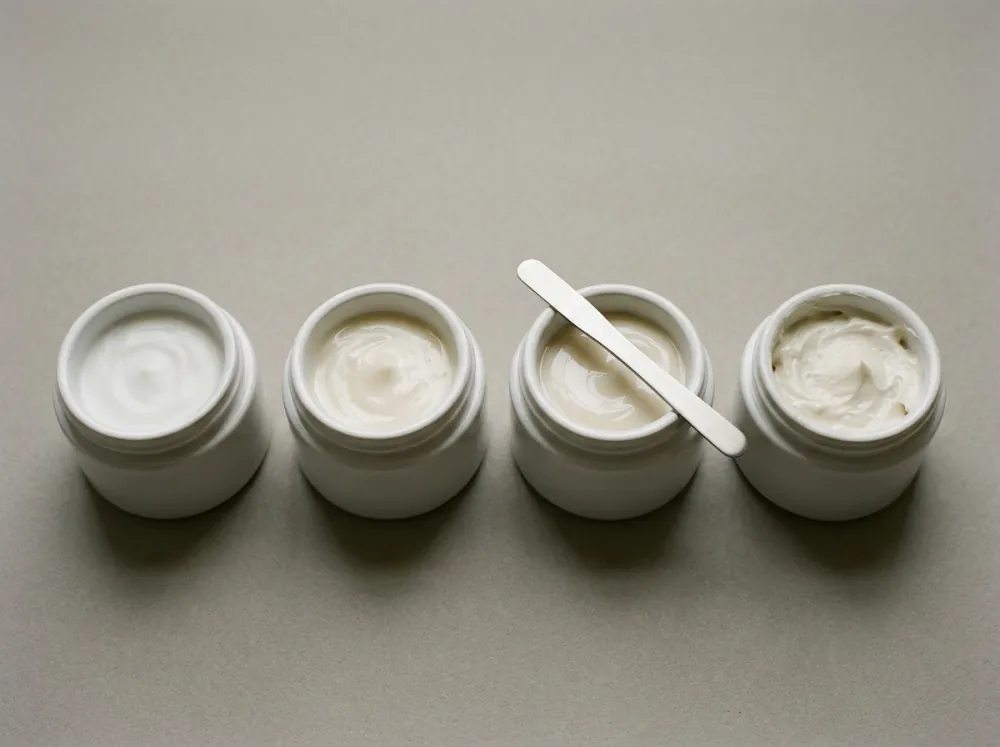 Four open white jars of cream in a row seen from slightly above on a grey-beige surface, the creams ranging from smooth white to thicker beige, with a spatula resting across the third jar.