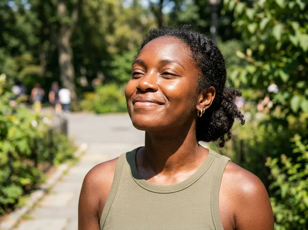 A woman with deep brown skin outdoors in bright sunlight, skin even-toned with no pale film, looking comfortable in the light.