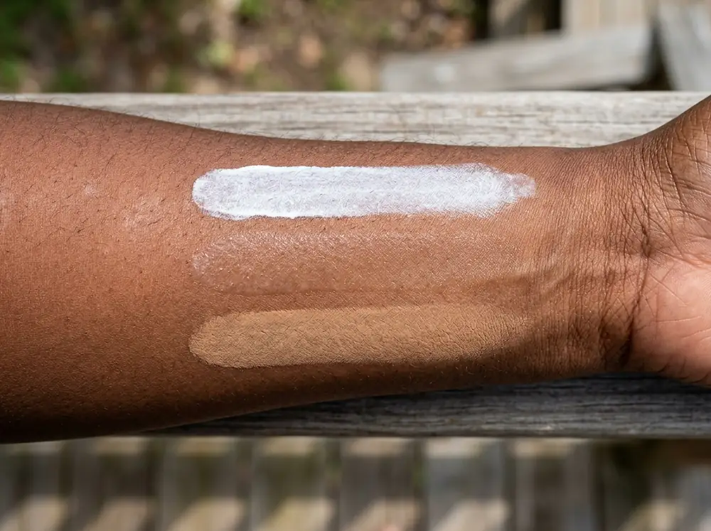 Three stripes of sunscreen swatched side by side on deep brown skin — one chalky white, one clear, one tinted and nearly invisible.