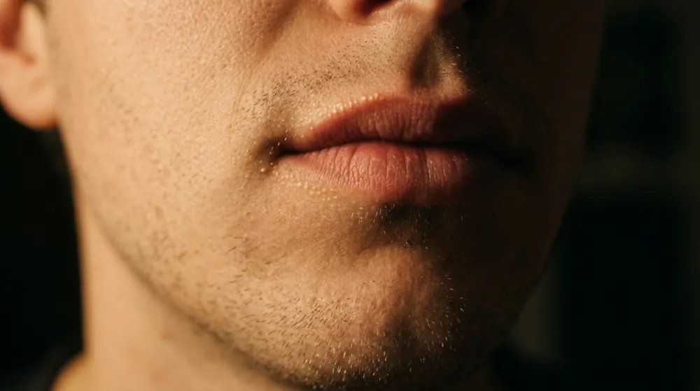 The lower half of a man's face in warm low lamp light against a dark background, lips closed, with fine stubble on the chin and tiny pale dots just visible along the upper lip border.
