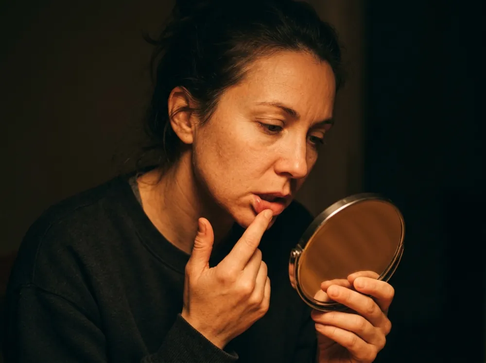 A woman in a dark room looking into a small round hand mirror while a finger draws her lower lip down to check its border, lit by a warm lamp.